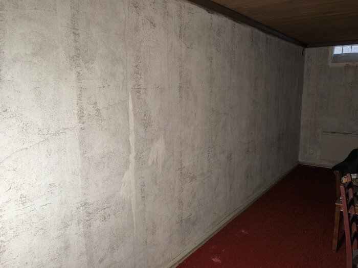 Betongvägg i gillestuga med ytliga sprickor, utan tapet och målad med plastfärg, mot en röd matta och möbler i ett gammalt suteränghus.
