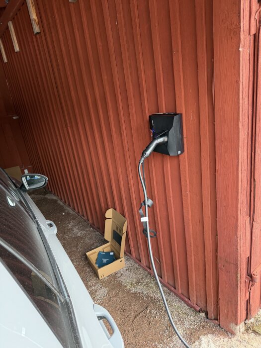 Laddbox installerad på röd trävägg med kabel kopplad till en bil. Kartong på marken vid sidan av bilen.
