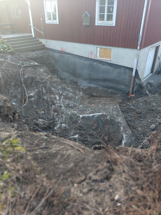 Grävd grop vid ett hus med kladdig jord och skyddande dämpmatta på husets grund.