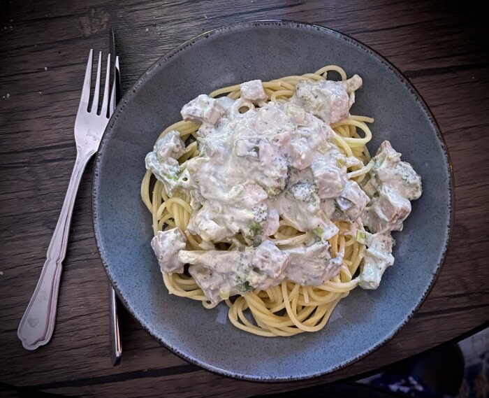Pastarätt med spagetti, pastasås innehållande fläskfilé, ädelost, broccoli, lök och gräddfil på en tallrik med en gaffel bredvid.