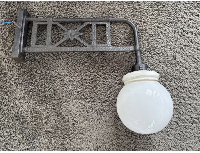 En antik vägglampa i smidesjärn med en vit rund glaskupa, placerad på en grå matta.