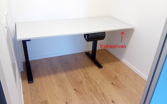 Höj- och sänkbart skrivbord i vit färg, med en tillagd extraskiva markerad med en röd pil, placerat mot en vit vägg.
