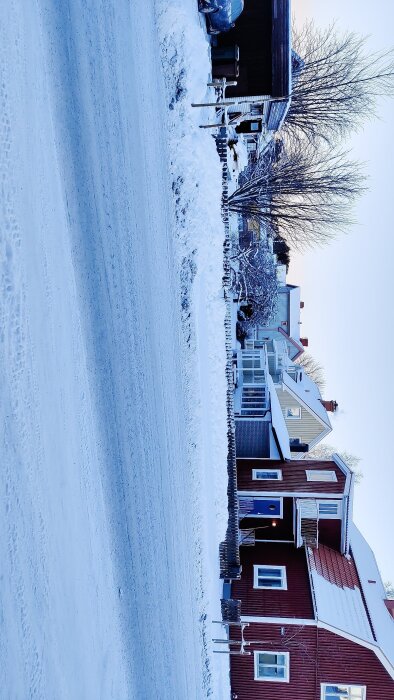 Gata med hus vid sidan, vintertid med snö täcker marken. Husen är röda med vita detaljer och står nära en väg där bilar kan passera.