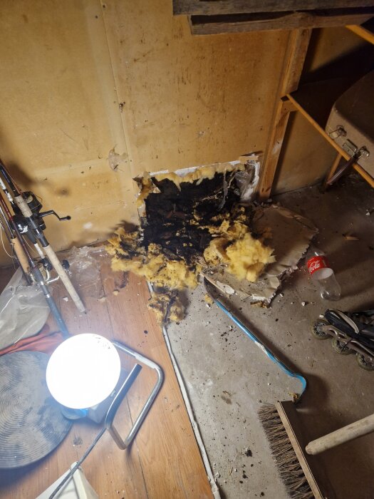 Skadad garagevägg med fuktskador där fasadpanelen börjat ruttna. Isoleringsmaterial syns på golvet, omringat av diverse verktyg och skräp.