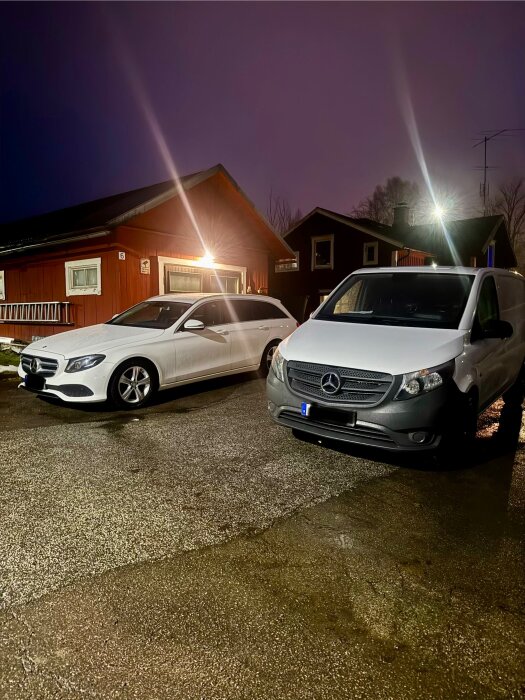 Två vita bilar parkerade framför ett rött hus på en våt kväll. Bilarna ser nytvättade ut.