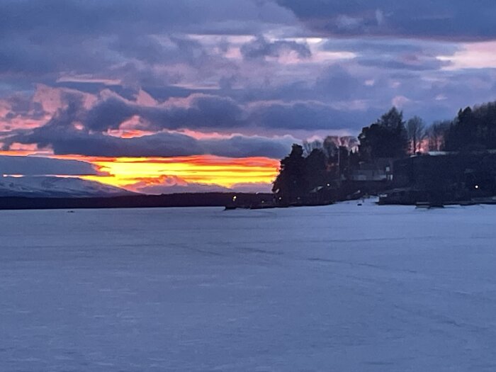 Solnedgång över ett fruset sjölandskap med träd och hus i silhuett mot en färgstark himmel.