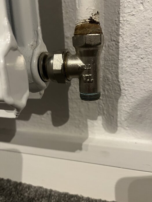 Närbild av en vit radiatoranslutning mot väggen, med en metallventil och ett delvis rostigt rör.