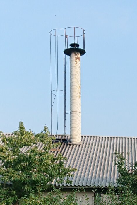 En vit skorsten med en stege och skyddsräcke på ett plåttak, omgiven av träd i förgrunden, mot en klarblå himmel.