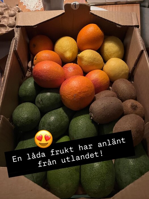 En låda med citrusfrukter, kiwi och avokado på ett bord. Text säger "En låda frukt har anlänt från utlandet!" med hjärtemoji.