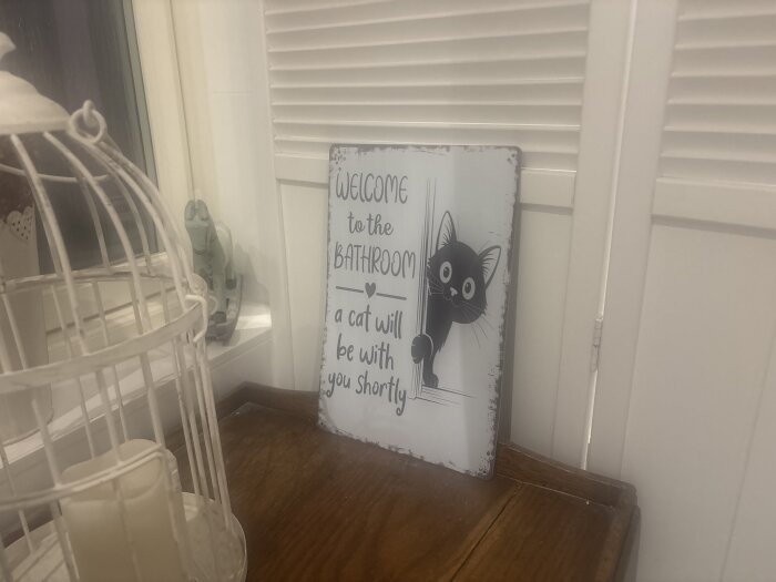 En skylt med en tecknad katt som tittar fram och texten "Welcome to the bathroom, a cat will be with you shortly", en fågelbur syns bredvid.