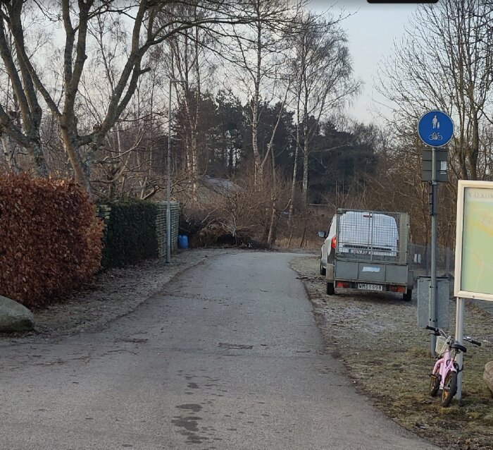 En smal cykelväg med ett fordon parkerat på, omgiven av träd och buskar, samt en vägskylt och en rosa barncykel vid sidan.