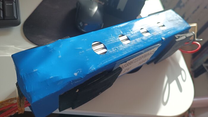 Blått rektangulärt cykelbatteri med synliga cirkulära öppningar och kablar, placerat på ett bord bredvid ett tangentbord och en datormus.