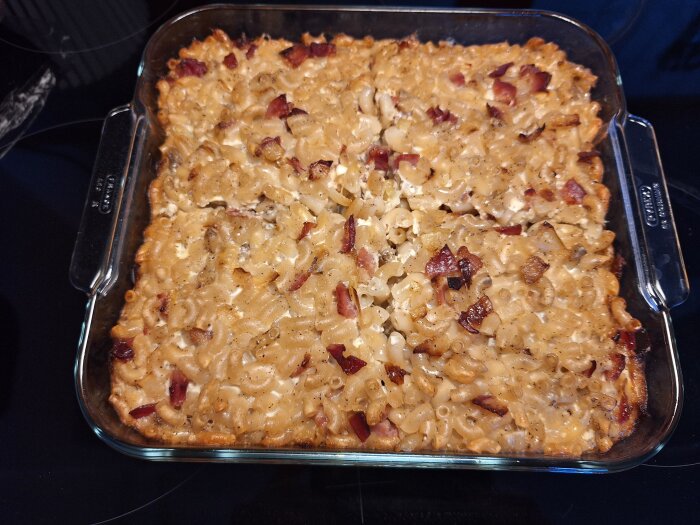 Ugnsform med makaronpudding med bitar av bacon eller skinka, stående på en spishäll.