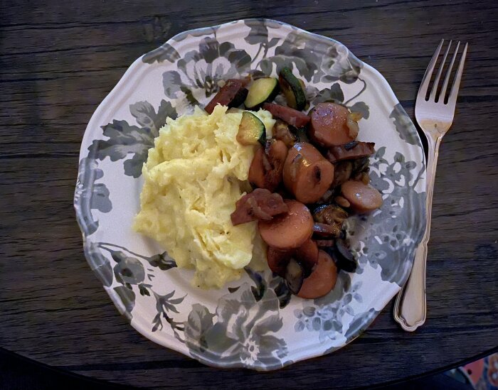 Tallrik med potatismos och stekta grönsaker med korvskivor, bredvid en gaffel på ett mörkt träbord.