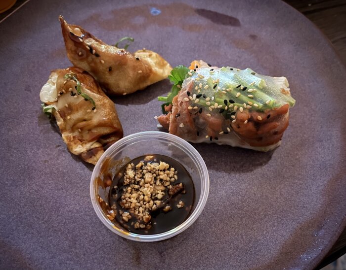 Vietnamesiska rätter på tallrik: två gyoza med anka, summerroll med fläsk, och en sås i liten plastkopp, dekorerade med sesamfrön.