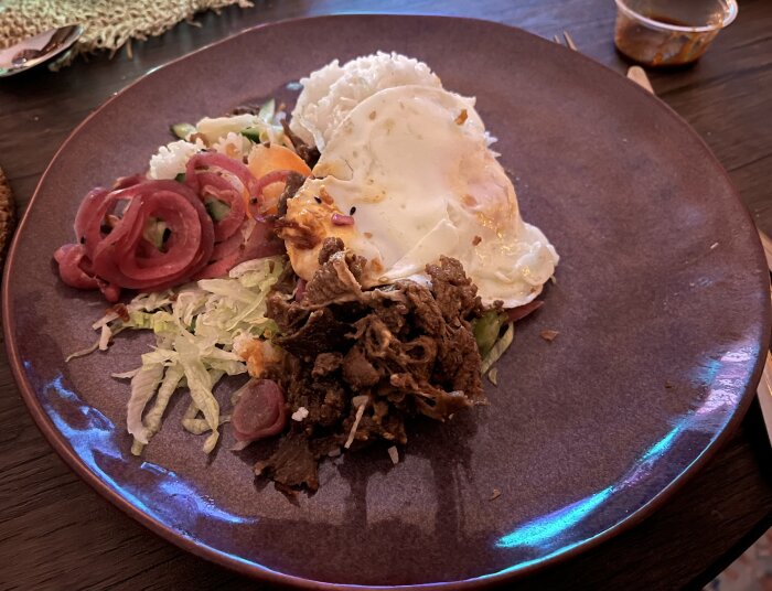 Tallrik med strimlat kött, stekt ägg, ris, picklad rödlök, sallad och grönsaker.