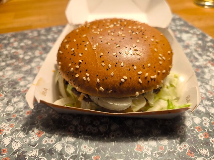 Hamburgare i förpackning med sesam- och vallmofrön på toppen, placerad på ett blommigt underlägg.