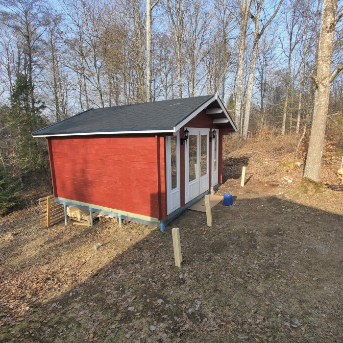 Rött träförråd med svart tak i skogsmiljö, beläget på en sluttning med träd runt omkring. Flera stolpar är uppställda runt byggnaden.