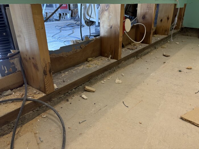 Bilden visar en väggkonstruktion i en källare där träsyllar och reglar står på en betongplatta, med byggmaterial och kablar runt omkring.