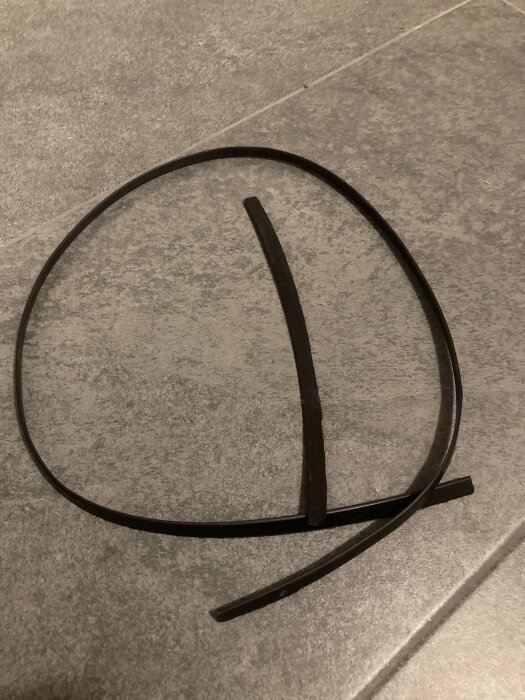 Trasig svart rem eller gummipackning på grått golv, cirka 60 cm lång.