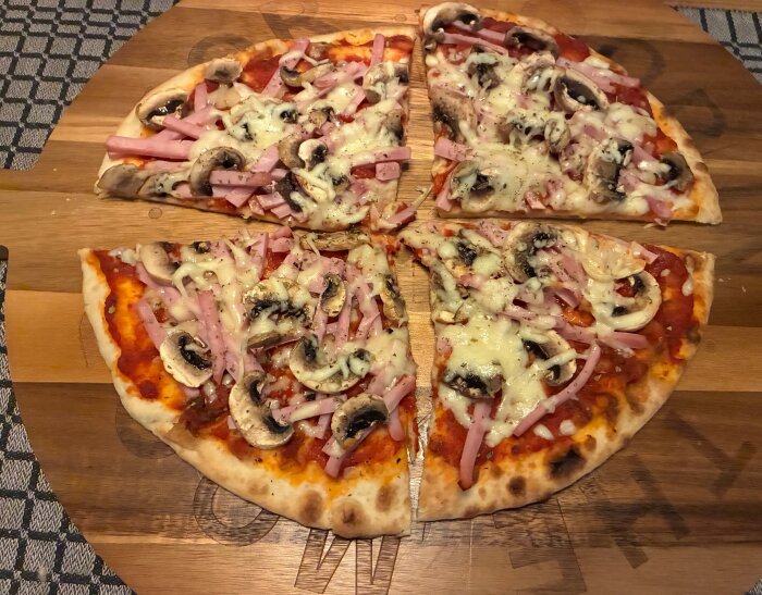 Pizza med mozzarella, skinka, färska champinjoner, Västerbottensost och grön Spetspaprika på träbricka. Fyra skivor syns ovanifrån.