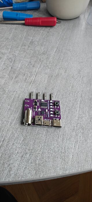Lila kretskort med flera USB-portar och komponenter på ett grått bord, omgiven av skruvmejslar och en vit kopp.