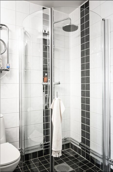 Duschhörna med glasdörrar, hörndusch med takdusch och handduk hängande på dörrhandtaget i ett helkaklat badrum.