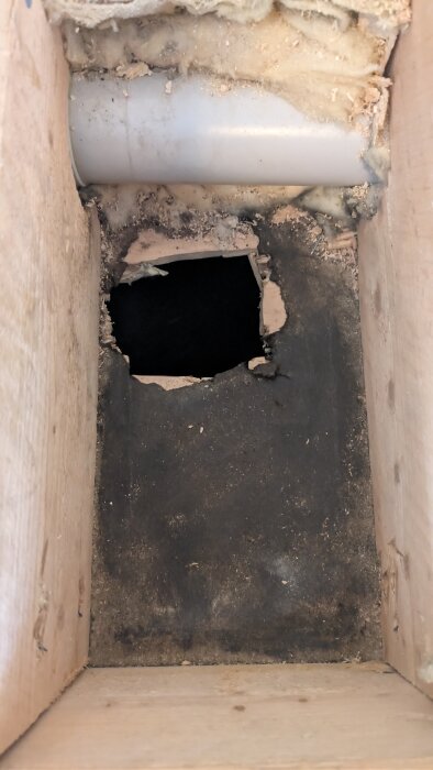 Hål i blindbotten i träbjälklag, med synlig isolering och en vit rörledning ovanför hålet.