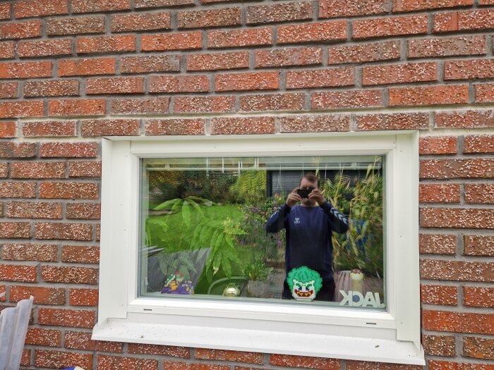 Spegelbild av en person som tar foto av ett fönster på en röd tegelvägg, med gröna växter i bakgrunden och en dekorativ figur i fönstret.