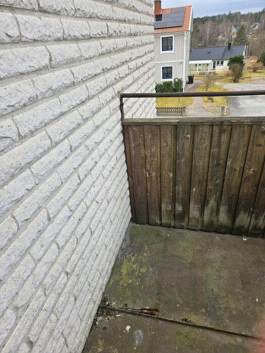 En äldre balkong med plåtgolv intill en tegelvägg, sett från en byggnad med trähus i bakgrunden och en vårdslösning.