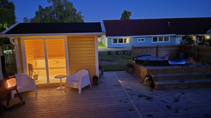 Belyst terrass med bastu i trä och spabad, omgiven av staket och vegetation, och hus i bakgrunden.