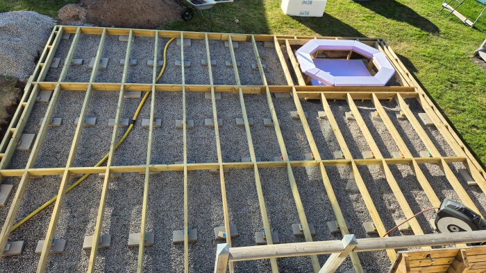 Träram för terrassgolv med grus och isolering placerad för badtunna. Gult flexibelt rör syns bland underredet. Utflyttning av terrass pågår.