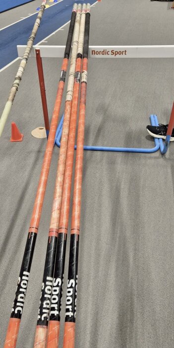 Stänger för stavhopp placerade på ett ställ bredvid en löparbana inomhus, märkta med "Nordic Sport".