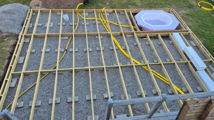 Byggarbetsplats för utvidgning av trädäck med trästomme, isolering, gula slangar för installation av spabad och bastu, synliga styroxlådor.