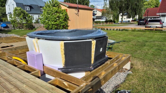 Isolerad spabadstunna med plasta, halvt inbyggd i trädäck med frigolitlådorna synliga för slangisolering, omgivet av en grönskande trädgård.