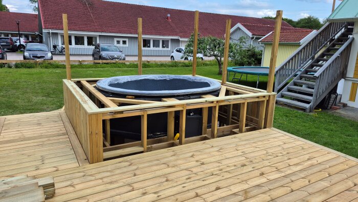 Bygge av spabad/terasspool inbyggd i trädäck med träram, omgiven av gräsmatta och trappa. Huset och bilar syns i bakgrunden.