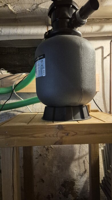 Sandfilterpump för spabad på en träram i källare, gröna slangar syns i bakgrunden.