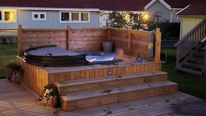 Spabad med träbeklädnad och lock på en terrass omgiven av träplank, kvällsbelysning och några växter i krukor.