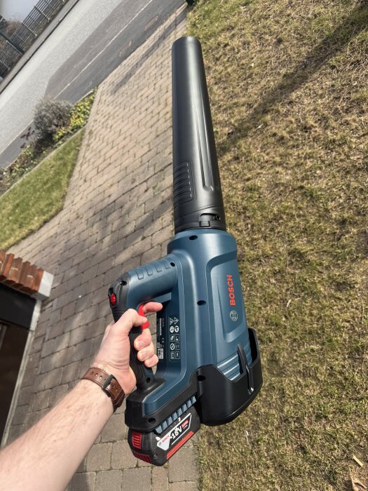 En person håller i en blå Bosch batteridriven lövblåsare utomhus på en stenlagd gångväg.