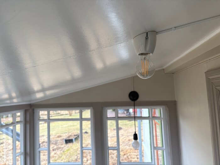 Taklampa med glödlampa i ett rum med stora fönster och utsikt över en gräsbevuxen gård.