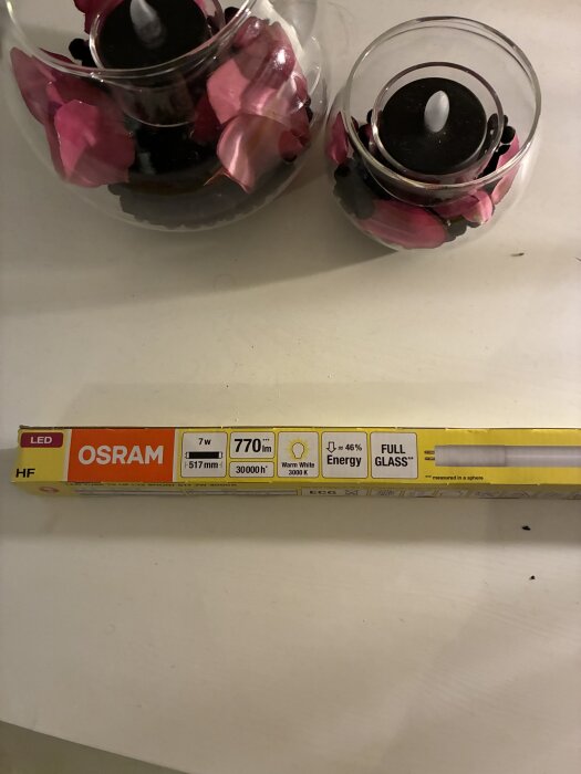 Förpackning av Osram LED-lysrör, 7W, 770lm, 517mm, varmvit 3000K, ligger på ett bord intill ljusdekorationer med rosa blad.