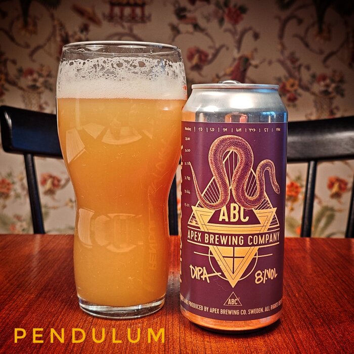 Ett glas med skummande öl bredvid en burk från Apex Brewing Company med lila och gul etikett på ett bord.