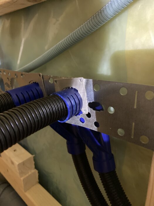 Installation av bock och PEX-rör med blå beslag på metallplatta mot grön bakgrund, trästrukturer synliga i nedre del.
