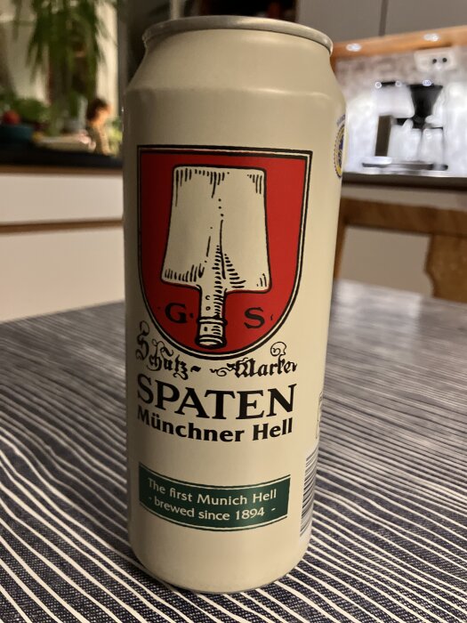 En burk Spaten Münchner Hell-öl står på ett randigt bord, med ett kök i bakgrunden.