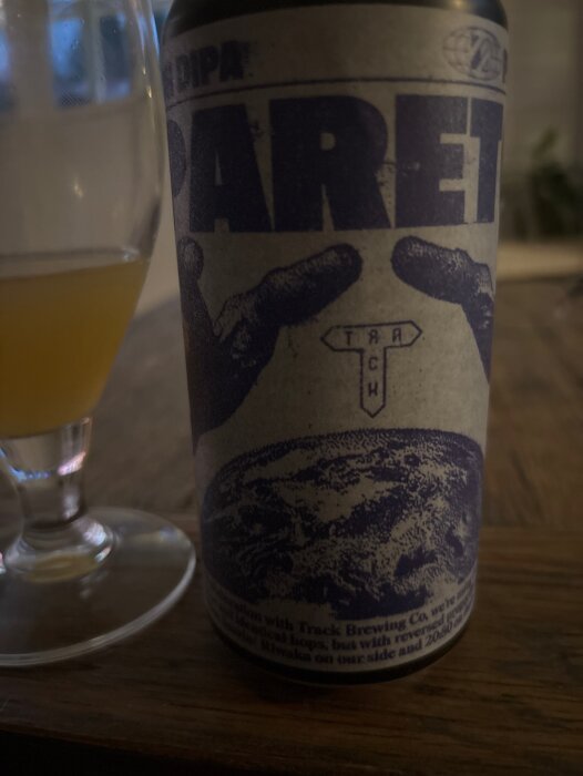 Ölglas och Pareto DIPA burk från Prizm Brewing med text och grafik, stående på ett bord i svagt ljus.