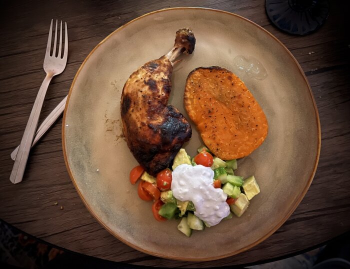 Grillad kycklingklubba med sötpotatis, sallad och ädelostkräm på en tallrik, med gaffel och kniv vid sidan.