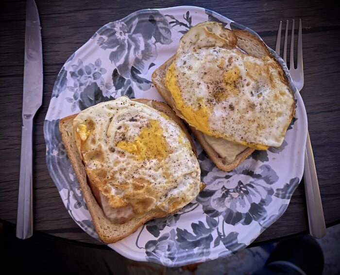 Två varma mackor med stekt ägg på en blommig tallrik, serverade med kniv och gaffel.