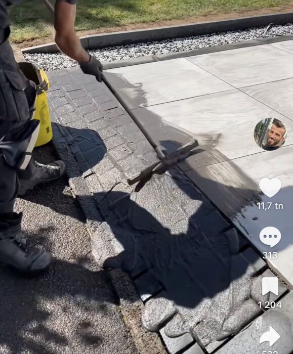 Person som applicerar hårdfog mellan stenar på en uppfart med hjälp av ett verktyg. Vit stenläggning och grus syns i bakgrunden.