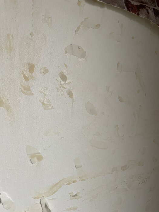 Närbild av vägg med kvarvarande tapetlim och fläckar efter borttagen plastig tapet, ojämn yta och synligt bakgrundslager av tapet.