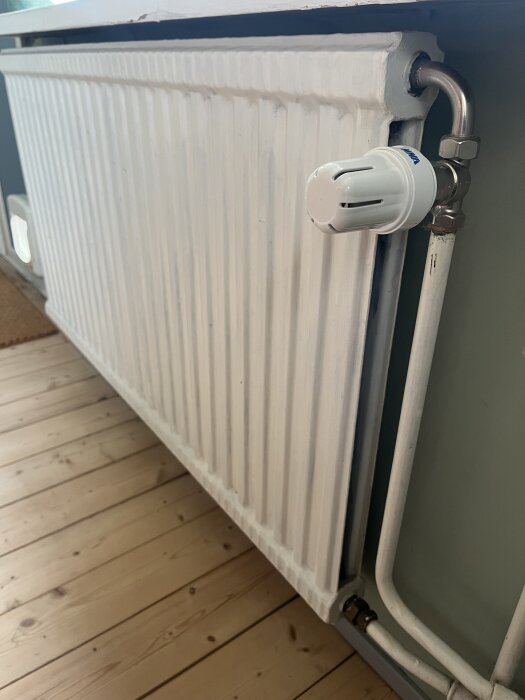 Ett vitt element med termostatventil på höger sida, monterat bredvid en trävägg i ett rum med trägolv.
